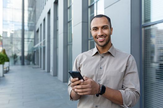 Portrait of African American businessman, man holding phone smiling and looking at camera, office worker in shirt outside modern office building on lunch break.
