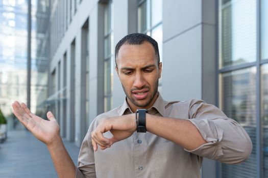 A man is looking unsatisfied at a smartwatch. A Latin American businessman is waiting outside an office building.