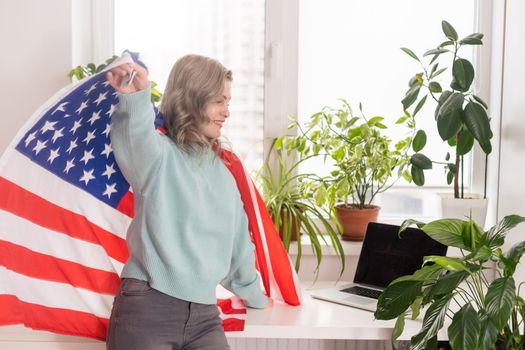 Happy woman employee sitting wrapped in USA flag, shouting for joy in office workplace, celebrating labor day or US Independence day