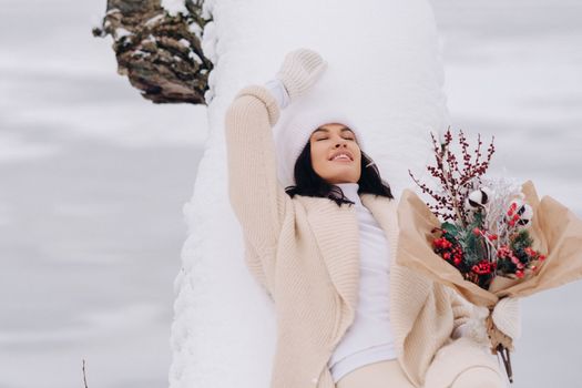 A beautiful girl in a beige cardigan and a white hat with flowers enjoys a snowy winter in nature.