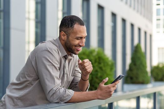Happy african american businessman looking at smartphone screen, man in shirt outside modern office building received good news of win and success online, boss celebrating financial triumph.