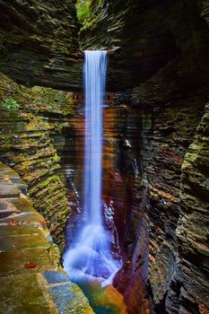 Image of Tall waterfall pours into deep gorge of layered rocks and path leading behind