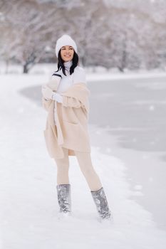 A beautiful girl in a beige cardigan and a white hat with a glass of tea enjoys a snowy embankment by the lake.