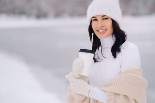 A beautiful girl in a beige cardigan and a white hat with a glass of tea enjoys a snowy embankment by the lake.