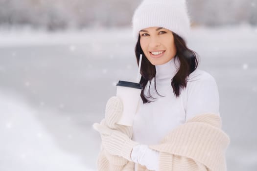 A beautiful girl in a beige cardigan and a white hat with a glass of tea enjoys a snowy embankment by the lake.