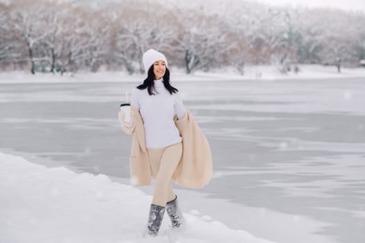 A beautiful girl in a beige cardigan and a white hat with a glass of tea enjoys a snowy embankment by the lake.