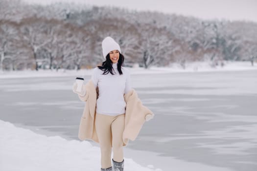 A beautiful girl in a beige cardigan and a white hat with a glass of tea enjoys a snowy embankment by the lake.