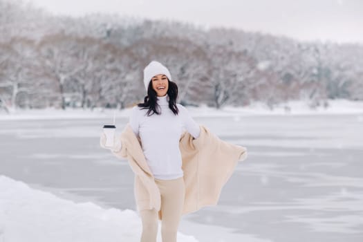A beautiful girl in a beige cardigan and a white hat with a glass of tea enjoys a snowy embankment by the lake.