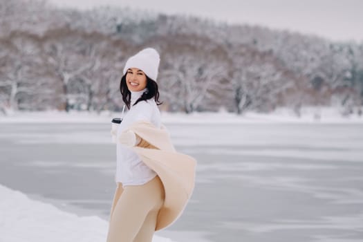 A beautiful girl in a beige cardigan and a white hat with a glass of tea enjoys a snowy embankment by the lake.