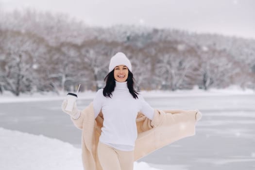 A beautiful girl in a beige cardigan and a white hat with a glass of tea enjoys a snowy embankment by the lake.