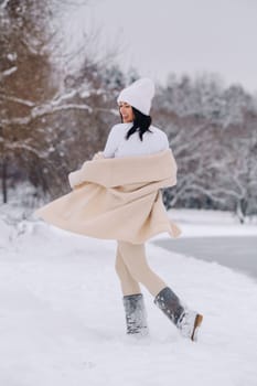 A beautiful girl in a beige cardigan and a white hat with a glass of tea enjoys a snowy embankment by the lake.