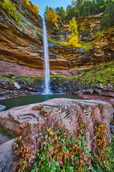 Image of Waterfall pouring over layered cliffs from below with rocks with red and green vines climbing up