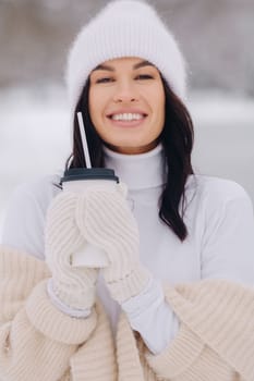 A beautiful girl in a beige cardigan and a white hat with a glass of tea enjoys a snowy embankment by the lake.