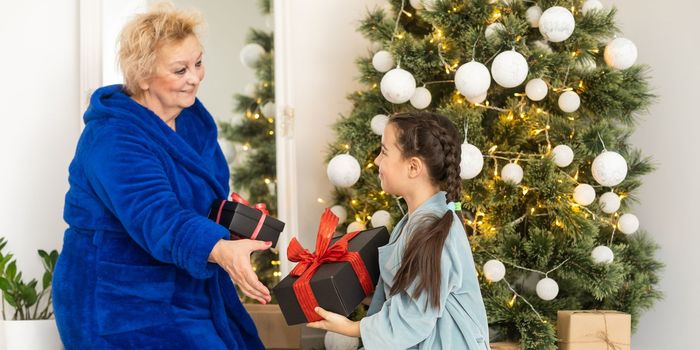 Cheerful grandma and her cute grand daughter girl exchanging gifts. Granny and little child having fun near tree indoors. Loving family with presents in room