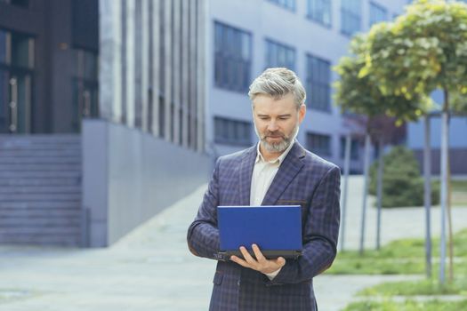 serious and concentrated gray haired boss working on laptop while standing outside modern office building, mature boss in business suit typing on netbook keyboard, senior successful businessman.