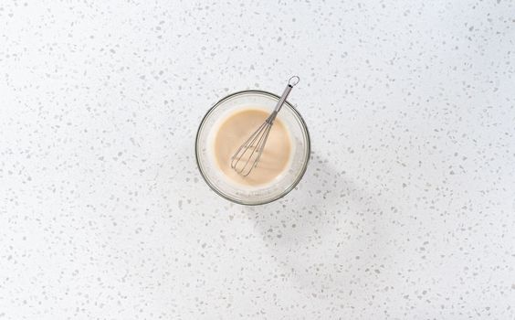 Flat lay. Mixing ingredients with a hand whisk in a glass mixing bowl to make the vanilla glaze.