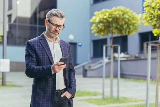 Serious and focused mature businessman using smartphone outside office building, gray haired man in glasses and business suit reading online message, senior boss walking in business city.