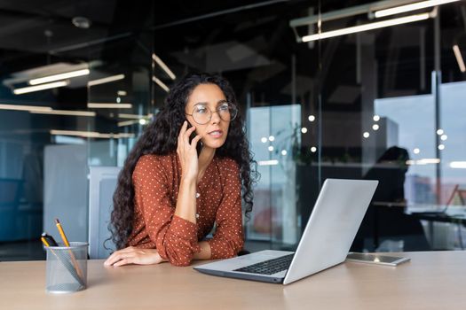 Young beautiful businesswoman talking on the phone and looking towards the window, happy hispanic woman inside the office using a laptop at work.