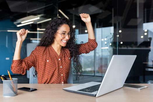 A beautiful young Latin American woman freelancer sits in the office at the table with a laptop, looks at the monitor, raised her hands, rejoices.