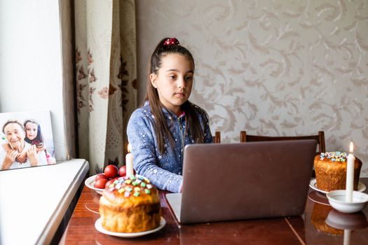 Pretty little girl painting easter eggs at home during coronavirus covid-19 outbreak. Kid using laptop on kitchen, online, video call to friends.