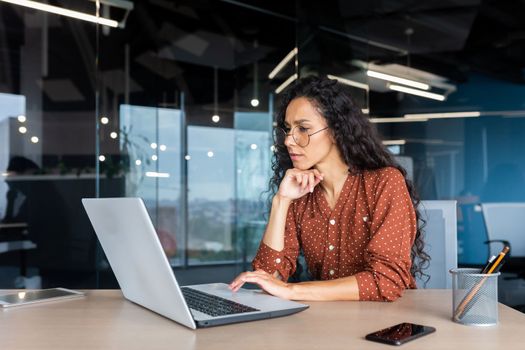 Serious thinking Latin American woman working inside office, businesswoman pondering complex decisions using laptop at work.