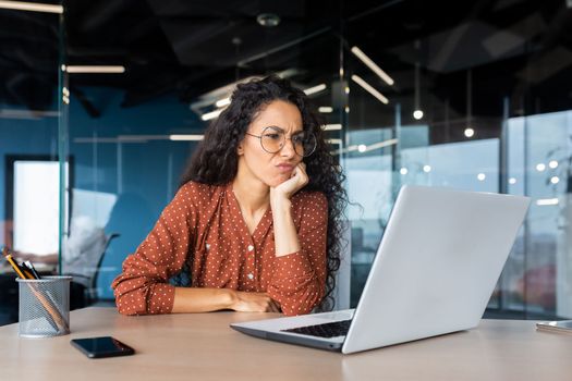 Serious thinking Latin American woman working inside office, businesswoman pondering complex decisions using laptop at work.