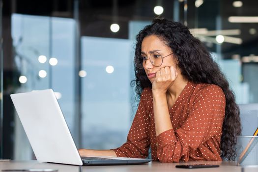 Serious thinking Latin American woman working inside office, businesswoman pondering complex decisions using laptop at work.