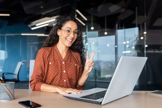 Cheerful and successful Latin American businesswoman talking remotely via video call with colleagues and partners, office worker inside the office using a laptop at work.