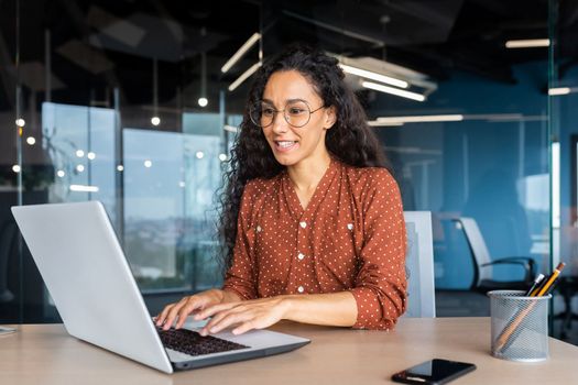 Happy and smiling hispanic businesswoman typing on laptop, office worker with curly hair and glasses happy with achievement results, at work inside office building.