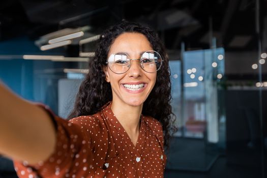 Business woman in the office talking on a video call, using application on smartphone for online remote communication, looking at the camera and smiling, a Latin American inside wearing glasses.