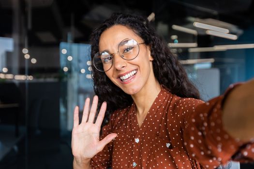 Business woman in the office talking on a video call, using application on smartphone for online remote communication, looking at the camera and smiling, a Latin American inside wearing glasses.