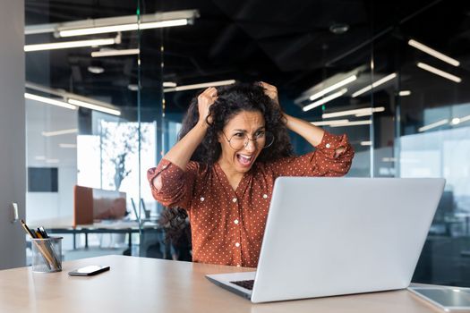 Upset woman at work yelling at laptop monitor, angry businesswoman holding hair on head inside office.