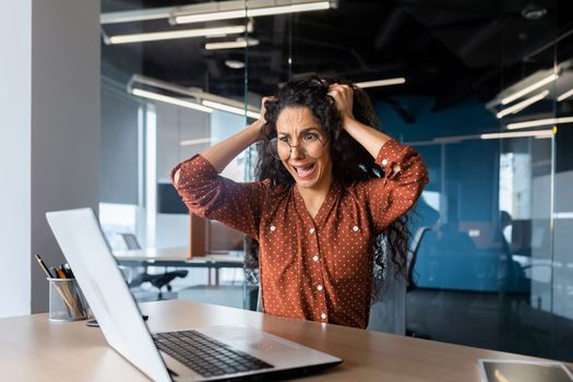 Upset woman at work yelling at laptop monitor, angry businesswoman holding hair on head inside office.