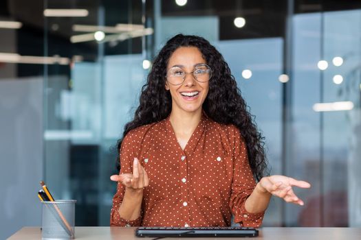 Online customer support video call, Latin American woman looking at web camera smiling and consulting customers, helpline worker working inside office.