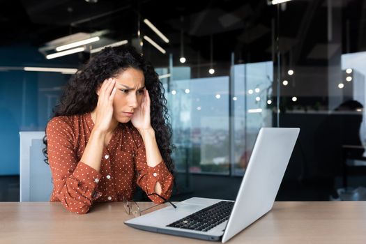 Overtired hispanic business woman working inside modern office, office worker with laptop having severe headache.