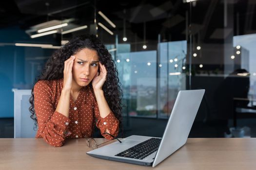 Overtired hispanic woman at work, business woman in glasses holding hands on head, having severe headache, working inside office sitting at table using laptop at work.