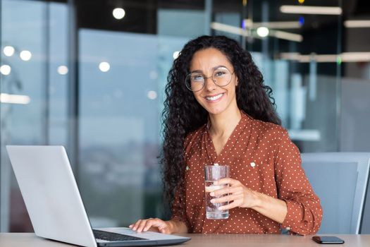 Portrait of Hispanic woman inside modern office at work, businesswoman smiling and looking at camera holding a glass of pure filtered water in her hand.