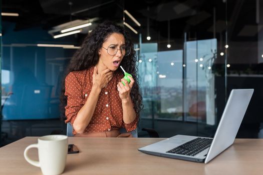 Sick woman in the office, Latin American business woman has a severe sore throat, uses medicine to relieve the pain.