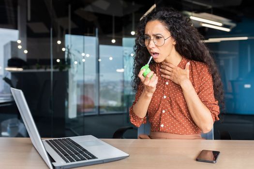 Sick hispanic woman in office, business woman has sore throat, sore throat, uses medicine for pain relief while sitting at work inside office with laptop at work.