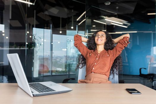 Happy and smiling successful hispanic business woman finished work satisfied with work result and achievement, hands behind head and smiling close up.