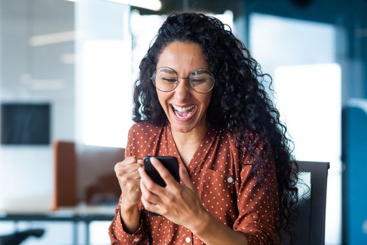 Happy and joyful hispanic business woman working inside office reading online message on phone, pleased and shocked celebrating victory success, close up photo of office worker in glasses.