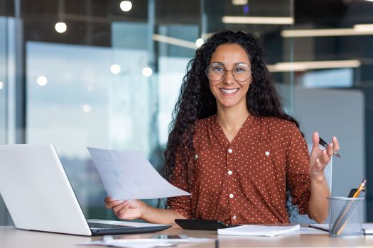 Portrait of happy and successful hispanic woman, businesswoman smiling and looking at camera holding contracts and invoices, working inside office with laptop on paper work.