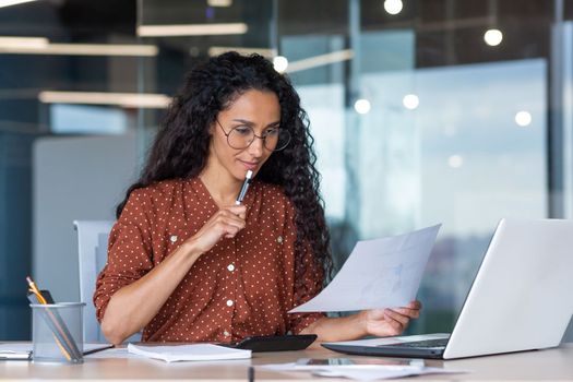 Beautiful hispanic woman in office doing paperwork, successful businesswoman financier working inside office sitting at table using laptop, at work with contracts and bills, thinking about decision.