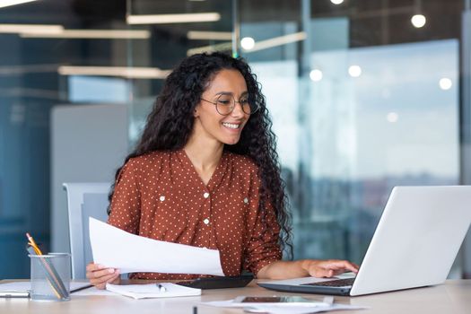 latin american businesswoman working inside office with documents and laptop, worker paperwork calculates financial indicators smiling and happy with success and results of achievement and work