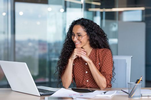 Young happy and successful businesswoman in glasses working with documents inside office, Hispanic woman with laptop looking at bills and contracts, financier with curly hair using laptop.
