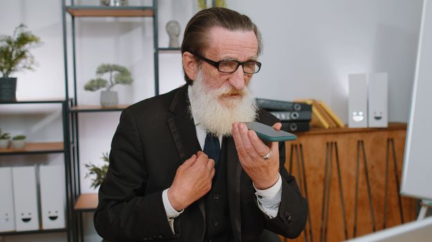 Senior old businessman working, having mobile phone loudspeaker talk at office desk with laptop. Elderly professional freelance man holding smartphone using messenger chat apps. Employment, occupation