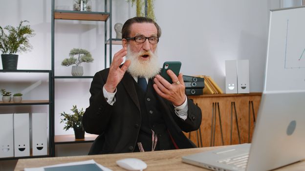 Oh my God Wow. Happy amazed senior businessman in suit working on smartphone shocked by sudden victory game winning lottery goal achievemen good news. Elderly grandfather man freelancer at home office