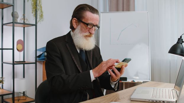 Elderly businessman programmer software developer working making online purchase payment shopping with credit bank card and smartphone at modern office desk with laptop. Senior freelancer business man