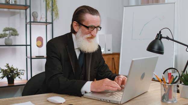 Senior businessman freelancer at office workplace, start working on laptop computer. Professional manager freelancer elderly man opens notebook, sends messages makes online purchases watching movies