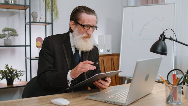Senior businessman working on laptop computer at home office desk talk on online communication video call with employee, boss. Manager grandfather old man freelancer support services looking at camera